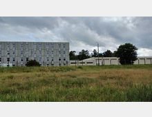 Ein graues, mehrstöckiges Betongebäude mit vielen symmetrisch angeordneten Fenstern neben einem flacheren weißen Gebäude, umgeben von einer Wiese im Vordergrund und Bäumen im Hintergrund unter einer bewölkten Himmel.