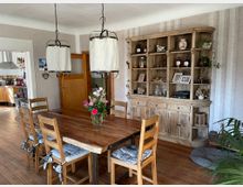 Esszimmer mit Holzboden, einem großen Holztisch und passenden Stühlen mit Sitzkissen. Im Hintergrund steht ein großer rustikaler Holzschrank mit offenen Regalfächern, in denen Deko-Objekte und Fotos stehen; links führt eine Tür in die angrenzende Küche.