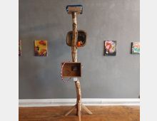 Ein Kunstobjekt aus Holz, bestehend aus einem vertikalen Stamm mit einer Plattform oben, einem geflochtenen Korb in der Mitte und einer Holzkiste sowie einem Hula-Hoop-Reifen unten, vor einer grauen Wand mit mehreren gemalten Bildern im Hintergrund.