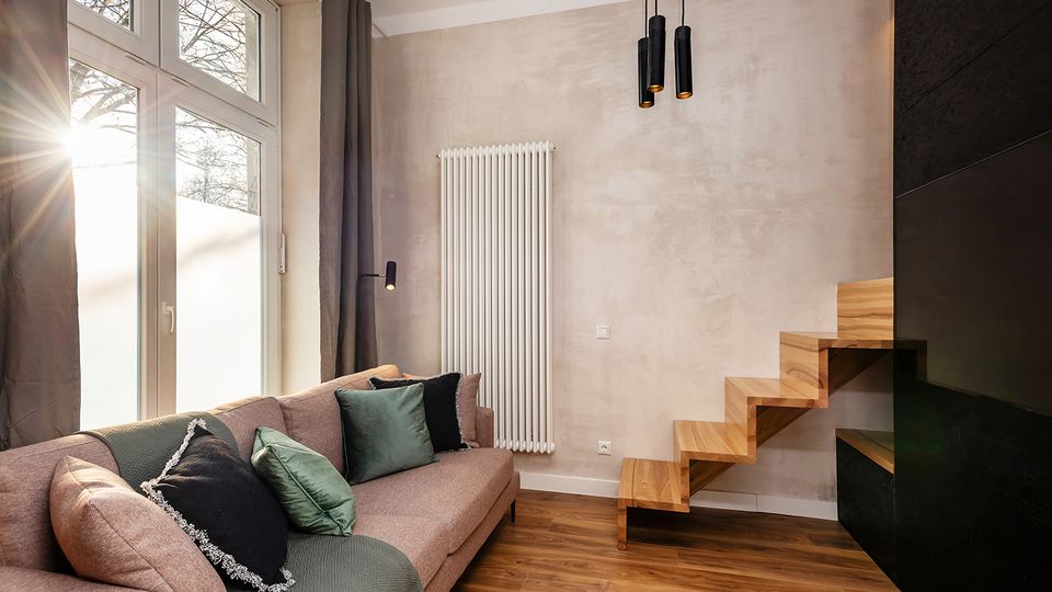 Modernes Wohnzimmer mit großem bodentiefem Fenster, grauen Vorhängen und Ecksofa mit Kissen auf Holzfußboden. An der Wand hängen ein hoher weißer Heizkörper und schwarze Pendelleuchten; rechts führt eine schwebende Holztreppe an einer dunklen Wand nach oben.