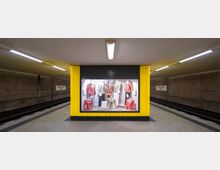 Eine U-Bahn-Station mit gelben Wandpaneelen und einer beleuchteten Schaufensterauslage in der Mitte, die Kleidung und Dekorationen präsentiert. Im Hintergrund sind die Bahnsteige und ein Schild mit der Aufschrift „Jakob-Kaiser-Platz“ zu sehen.