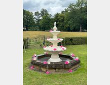 Weißer, mehrstöckiger Springbrunnen mit rosafarbenen Blumen dekoriert, umgeben von einem achteckigen steinernen Becken. Der Brunnen steht auf einer Rasenfläche in einer parkähnlichen Umgebung mit Bäumen und einem Metallzaun im Hintergrund.