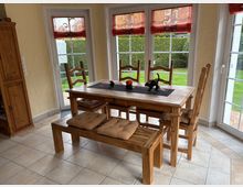 Essbereich mit Holzmöbeln in einem hellen Raum. Ein Holztisch ist von Stühlen und einer Bank mit Polsterauflagen umgeben, große Fenster mit roten Plissees bieten Blick auf eine grüne Hecke und einen Rasen.