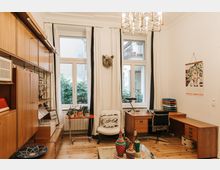 Innenraum eines gemütlich eingerichteten Arbeitszimmers mit Holzmöbeln. Der Raum hat große Fenster mit weißen Vorhängen, ein hölzernes Regal, einen Schreibtisch und eine Sitzgelegenheit; dekorative Elemente wie Pflanzen, bunte Körbe und eine Pop-Art-Wanddekoration prägen das Ambiente.