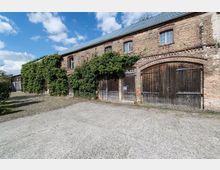 Backsteinbau mit mehreren Holztoren und kleinen Fenstern unter einem grauen Schrägdach, bedeckt von Rankpflanzen. Der Vordergrund zeigt einen gepflasterten Hof bei sonnigem Wetter.