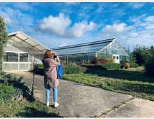 Eine Person steht auf einem gepflasterten Weg vor einem großen Gewächshaus mit gläsernem Dach und Wänden. Die Umgebung ist von Sträuchern, Hecken und grünem Rasen umgeben, unter einem teilweise bewölkten Himmel.