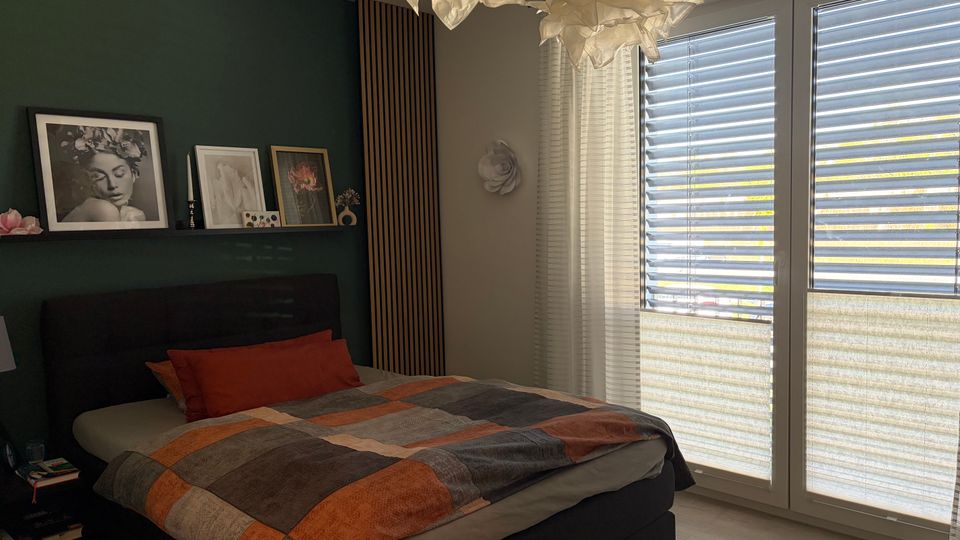 Modernes Schlafzimmer mit großem Polsterbett und karierter Bettdecke vor einer dunkelgrünen Akzentwand mit Bilderleiste. Rechts bodentiefe Fenster mit Lamellenjalousien und transparenten Vorhängen; an der Decke eine wolkenartige Hängeleuchte.