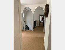 Flur in einem Wohnhaus mit hellem Teppichboden, weißen Wänden und mehreren Rundbögen. Auf der rechten Seite hängt eine Dekoration aus Zweigen, und links befindet sich ein Treppenabgang mit schwarzem Geländer.