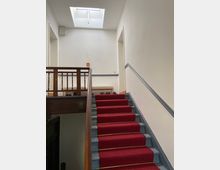 Treppenaufgang in einem Innenraum mit roten Teppichläufern auf den Stufen, blau gestrichenen Seitenleisten und einem Holzgeländer. Oben befindet sich eine helle Wand mit zwei offenen Türrahmen und einem Dachfenster, das Tageslicht hereinlässt.