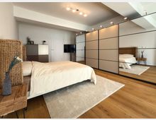 Modernes Schlafzimmer mit einem großen Bett mit beigen Bettbezügen, einem Nachttisch aus Holz und einer Vase darauf. Der Raum hat einen Holzfußboden, einen großen Spiegel an einem Einbauschrank und minimalistische Beleuchtung.