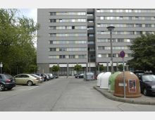 Ein mehrstöckiges Wohngebäude in moderner Architektur mit grauer Fassade, umgeben von parkenden Autos. Im Vordergrund befinden sich mehrere Glas- und Papiercontainer sowie einige Bäume und Straßenschilder.
