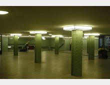 U-Bahn-Station mit grünen, genieteten Metallstützen, rechteckigen Deckenleuchten und grün gekachelten Wänden. Im Hintergrund sind eine Treppe und ein Fahrstuhl sowie Personen im Bereich sichtbar.