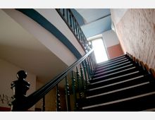 Treppenhaus mit einer schwarzen Holztreppe und dekorativ verzierten Geländern. Die Wände bestehen aus verputztem Mauerwerk in roten und weißen Farbtönen, oben ein Fenster, das Tageslicht hereinlässt.