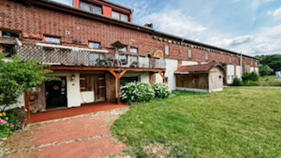 Langes Backsteingebäude mit kleinem Vorbau und Überdachung, davor ein roter Betonweg und ein großer Rasenbereich. Im Hintergrund sind ein blauer Himmel, Bäume und weitere niedrige Gebäudeteile zu sehen.