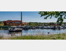 Kleiner Hafen mit anlegenden Motor- und Segelbooten an einer ruhigen Wasserfläche, im Hintergrund ein modernes rotes Gebäude mit Glasfronten und einer orangefarbenen Wasserrutsche, umgeben von Grünflächen und Bäumen. Ein hoher Schornstein ragt über das Gebäude hinaus, daneben sind Wohnhäuser und landschaftliche Hügel sichtbar.
