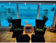 Wohnbereich mit zwei grünen Sessel im Vordergrund, einem kleinen Holztisch und Blick durch eine große Glasfront auf einen Innenpool. Eine mediterrane Wandmalerei mit Balustrade und Landschaft ergänzt die Poolumgebung.