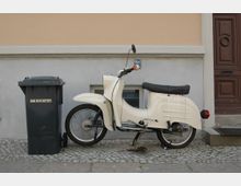 Ein cremefarbenes, klassisches Moped steht auf einem Gehweg vor einer beigefarbenen Hauswand. Daneben befindet sich eine schwarze Mülltonne, und rechts ist ein braunes Holztürportal erkennbar.