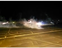 Eine nächtliche Flughafenlandschaft mit beleuchteten Start- und Landebahnen, Rauchentwicklung und einem beleuchteten Bereich im Hintergrund, in dem ein Flugzeug sichtbar ist. Weitere Flugzeuge und Lichter sind im Hintergrund zu erkennen.