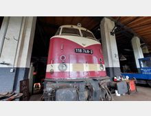 Eine rote und cremefarbene historische Diesellokomotive (Modellnummer 118 748-3) steht in einer alten Lokomotiv-Werkhalle mit Backsteinwänden und Holzbalkendecke, flankiert von Werkstattutensilien und einem blauen Waggon.