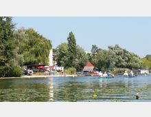 Ein Flussufer mit dichter Begrünung, einem kleinen Sandstrand und mehreren Sonnenschirmen. Im Vordergrund befinden sich Personen in Kanus auf dem Wasser, das mit Seerosen bedeckt ist; im Hintergrund stehen Gebäude und ein Steg.