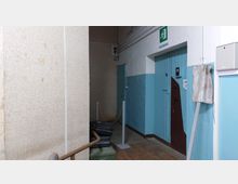 Ein schmaler Flur mit blau-weiß gestrichenen Wänden, einer Holztür mit einem "Ausgang"-Schild darüber und einem Sicherheitsgriff. Im Vordergrund stehen eine Metallstange und mehrere abgedeckte Gegenstände.