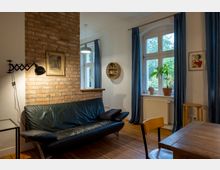 Wohnzimmer mit einem Sofa aus schwarzem Leder vor einer Wand aus Ziegelsteinen, an der ein ausziehbarer Wandstrahler befestigt ist. Rechts befinden sich zwei Fenster mit blauen Vorhängen, eine Fensterbank mit Topfpflanze, und ein Holztisch mit Stühlen auf einem Holzboden.