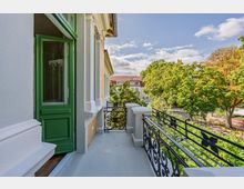 Balkon an einer historischen Fassade mit weiß verzierten Stuckelementen, grüner Balkontür und schwarzem schmiedeeisernen Geländer. Blick über hohe Laubbäume auf ein gegenüberliegendes Gebäude mit rotem Dach unter blauem Himmel.