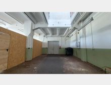 Ein leerer, industriell wirkender Raum mit weiß gestrichenen Wänden, teilweise grüner Sockelfarbe und einer Decke mit sichtbaren Trägern. Die Rückwand zeigt ein verschlossenes Doppeltor und eine aufgestellte Mülltonne, während links Holzverkleidungen und eine Tür sichtbar sind.