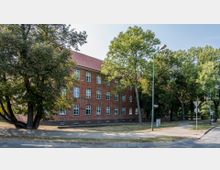 Backstein-Gebäude mit roten Ziegelfassaden und weißen Fenstern, umgeben von hohen Bäumen und einer Grünfläche. Im Vordergrund führt ein Gehweg entlang einer Straßenecke mit Laternen und Verkehrsschildern.