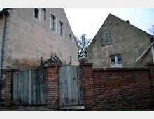 Zwei ältere Gebäude mit verputzten, verwitterten Fassaden und kleinen, einfach verglasten Fenstern, getrennt durch eine rote Ziegelmauer mit einem hölzernen Tor; im Hintergrund sind kahle Äste eines Baumes sichtbar.