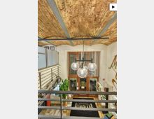 Blick von einer Empore in einen kleinen Laden mit freiliegender Ziegeltonnen-Decke und dunklen Stahlträgern. Unten sind ein Eingang mit vertikalen Lamellen, eine dreiflammige Kugel-Hängeleuchte, Metallgeländer sowie Vitrinen und Pflanzen im Holzfußboden-Bereich zu sehen.