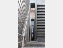 Blick in ein vertikales Treppenhaus mit grauen Steintritten und einem rechteckigen offenen Schacht in der Mitte. Die weißen, senkrechten Geländerstäbe rahmen die Treppe ein, die über mehrere Etagen hinabführt.