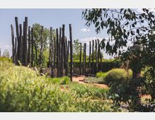 Holzskulpturen stehen in einer Landschaftsgestaltung mit grünen Sträuchern, Gräsern und Wegen aus rotem Kies, umgeben von Bäumen und blauer Himmel im Hintergrund.