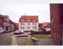 Kleiner Platz mit Kopfsteinpflaster, umgeben von historischen Gebäuden mit roten Ziegeldächern. Im Hintergrund steht ein weißes Haus mit drei Etagen, daneben sind weitere Fachwerkhäuser sichtbar.