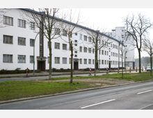 Weiße Wohngebäude im Bauhausstil mit flachen Dächern und gleichmäßig angeordneten rechteckigen Fenstern entlang einer Straße, auf der Baumschutzmaßnahmen und Verkehrsbeschränkungen zu sehen sind. Im Vordergrund befinden sich eine Straße und kleine Grünflächen mit einzelnen, kahlen Bäumen.
