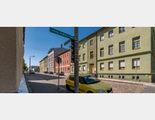 Straßenszene mit Kopfsteinpflaster, flankiert von mehrstöckigen Wohngebäuden in verschiedenen Farben, darunter ein grünes Gebäude im Vordergrund. Ein Straßenschild mit der Aufschrift „Ludwig-Sandberg-Straße“ ist sichtbar, und geparkte Autos stehen entlang der Straße, darunter ein gelbes Auto im Vordergrund.