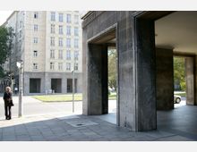 Straßenansicht mit einem hohen Gebäude im historischen Stil, das helle Fassaden mit zahlreichen Fenstern hat. Im Vordergrund sind massive dunkle Säulen eines Durchgangs sowie ein Bürgersteig und eine Ampel sichtbar, im Hintergrund eine Grünfläche mit Bäumen.