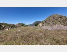 Weite Fläche mit trockenem Gras und mehreren Kieshügeln, teilweise mit Gras und Sträuchern bewachsen, unter einem klaren blauen Himmel. Im Hintergrund sind kleinere Büsche und Bäume sichtbar.