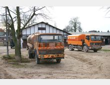 Zwei Tanklastwagen auf einem sandigen Hof vor einem großen Fachwerkgebäude mit weißer Fassade und dunklen Balken, umgeben von kahlen Bäumen und weiteren Gebäuden im Hintergrund.