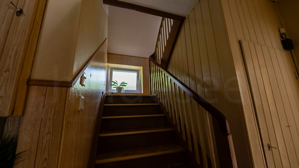 Innenaufnahme eines schmalen Treppenhauses mit Holzvertäfelung und dunkel gestrichener Holztreppe mit Geländer, die zu einem Podest führt. Am Ende des Treppenlaufs befindet sich ein Fenster mit einer Topfpflanze auf der Fensterbank, seitlich sind Türen und eine Wandleuchte zu sehen.