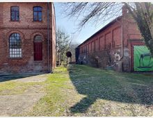 Zwei historische Backsteingebäude, eines links mit hohen Fenstern und roter Tür, das andere rechts mit einer langen Fassade und teilweise gemauertem Sockel. Im Vordergrund eine Grasfläche, im Hintergrund Bäume und ein Plakat mit einer schwarzen Pferdefigur.