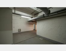 Kellerraum mit weißen Ziegelwänden, Betonboden und sichtbaren Lüftungsrohren an der Decke. Der Raum ist spärlich beleuchtet und hat eine Türöffnung, die in einen angrenzenden Bereich führt.