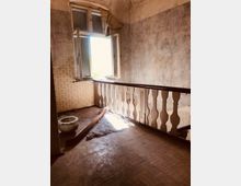 Ein verlassener Innenraum mit abgenutzten Wänden und einem offenen Fenster, durch das Tageslicht einfällt. Im Vordergrund ein hölzernes Geländer und auf dem Boden liegen Ablagerungen, darunter ein Stoffstück und Teile eines zerkleinerten Gegenstands.