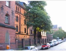 Straßenansicht mit einem historischen Backsteingebäude, das mehrere Stockwerke und kunstvolle Rundbogenfenster aufweist. Im Vordergrund stehen Bäume entlang des Gehwegs und an der Kopfsteinpflasterstraße sind parkende Autos zu sehen.