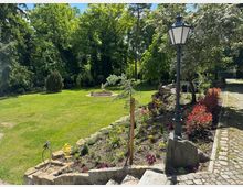 Gepflegter Garten mit einer grünen Rasenfläche, gesäumt von Bäumen und blühenden Sträuchern. Im Vordergrund ein gepflasterter Weg mit einer schwarzen Laterne und einem Blumenbeet, im Hintergrund eine steinerne Statue inmitten eines kleinen Brunnens.