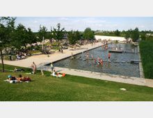 Ein belebter Park mit einem großen flachen Wasserbecken, wo Kinder und Erwachsene spielen. Im Vordergrund liegt eine Rasenfläche mit entspannenden Besuchern, während weitere Spielbereiche mit Schaukeln und schattenspendenden Bäumen im Hintergrund zu sehen sind.