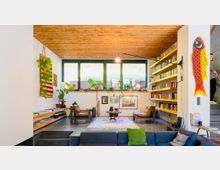 Wohnzimmer in modernem Design mit Holzbalkendecke, großen Fenstern und minimalistischem Mobiliar. Links hängen Pflanzen und eine dekorative Wandmatte, rechts befindet sich ein großes Bücherregal, dazwischen zwei Ledersessel und ein kleiner Tisch auf einem Teppich.