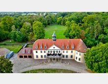 Luftaufnahme eines historischen Herrenhauses mit beigefarbener Fassade und rotem Ziegeldach, umgeben von einem großen Park mit dichtem Baumbestand. Im Vordergrund befinden sich eine Auffahrt mit Kiesbelag und ein angebautes Nebengebäude.