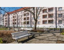 Wohngebäude mit pastellrosafarbener Fassade und roten Ziegeldächern, im Vordergrund ein kleiner Parkbereich mit weißen Parkbänken, gepflasterten Wegen und kahlen Bäumen. Ein Zaun trennt den Park vom Gebäude.