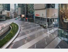 Moderne Innenhofszene mit Glas- und Metallfassaden umgeben von Geschäften, darunter ein Starbucks-Café. Im Vordergrund geschwungene Grünflächen über einer unterirdischen Ebene, die durch Glasabdeckungen sichtbar ist, und ein gepflasterter Platz mit integrierten Lichtelementen.